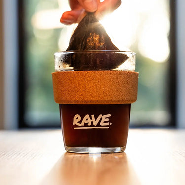 A hand lifts a used Colombia El Carmen Compostable Coffee Bag from a glass filled with dark coffee. The glass, featuring a cork grip and RAVE COFFEE branding, sits in sunlight streaming through windows in the background—highlighting this convenient way to enjoy specialty coffee at home.