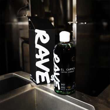 A bottle of RAVE COFFEE Coffee Liqueur stands on a metal countertop next to its black box, both with bold white text, set against a blurred industrial background.