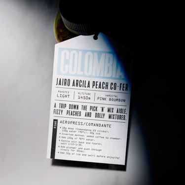A coffee label for RAVE COFFEE’s #0005 Colombia Jairo Arcila Peach Co-ferment - 150g displays information about its light roast, 1450m altitude, Pink Bourbon varietal, and includes Aeropress brewing instructions. Attached to a jar, the tag highlights this uniquely peach co-fermented specialty Colombian coffee as a standout offering.