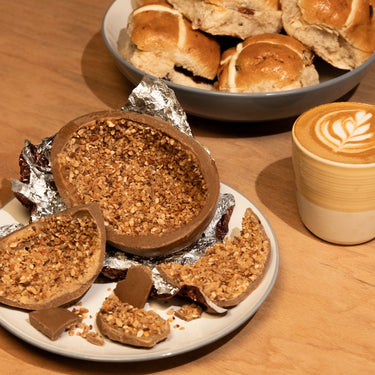 A halved RAVE COFFEE Coffee Chocolate Easter Egg filled with caramelised hazelnuts sits beside a latte with art, while hot cross buns rest in a bowl on a wooden table.