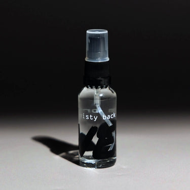 A small RAVE COFFEE RDT Bottle (I'm Bringing Misty Back), perfect for the Ross Droplet Technique to reduce coffee static, stands on a gray surface under a spotlight with its black and white label visible against a dark background.
