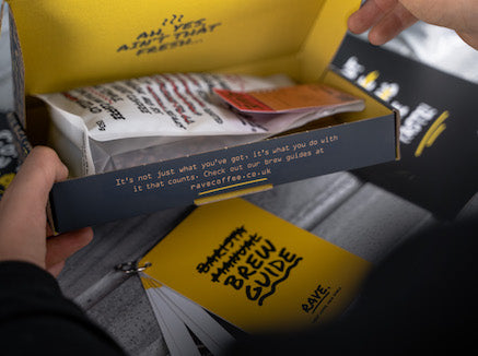 A person opens a yellow box from the Discovery 6 Month Gift Subscription, revealing a coffee packet and a BREW GUIDE booklet. Branded text appears on the inside flap and lid, with the BREW GUIDE visible on the table.